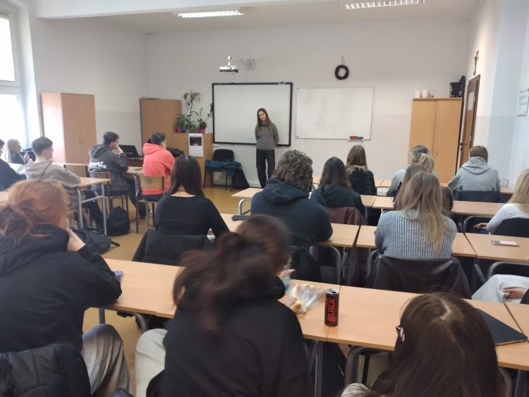 młodzież siedząca w szkolnych ławkach słucha wykładu kobiety