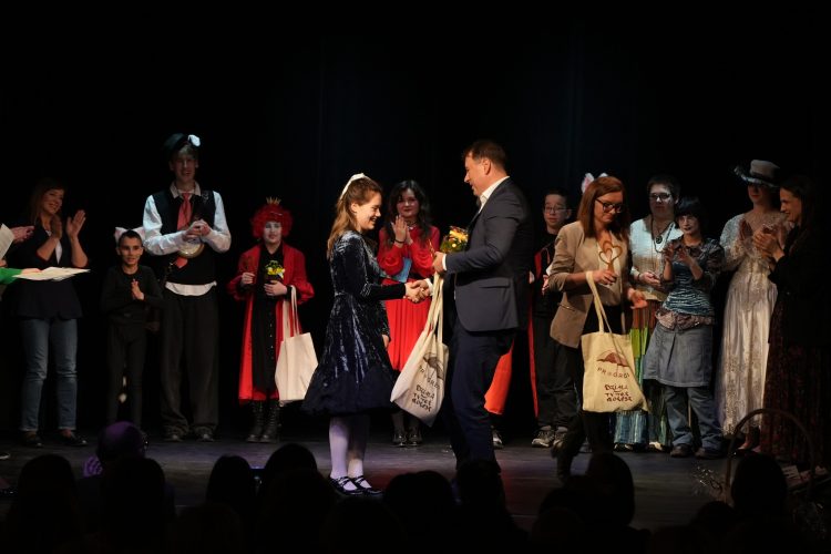 scena na niej aktorzy i dziewczynka otrzymująca nagrodę od mężczyzny w marynarce