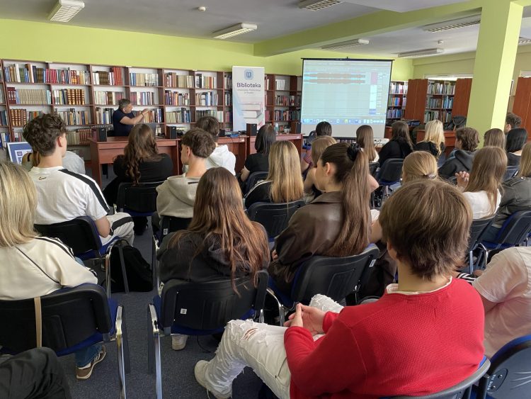 Uczniowie siedzą na krzesłach w sali bibliotecznej podczas warsztatów dotyczących programu do edycji dźwięku