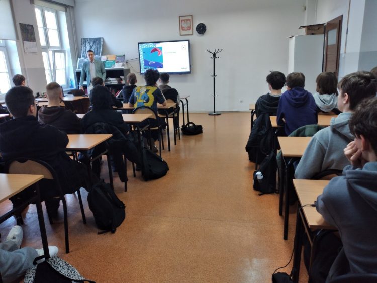 Sala lekcyjna, w której młodzież siedzi przy ławkach zwrócona w stronę prelegenta stojącego przy monitorze interaktywnym z wyświetloną prezentacją