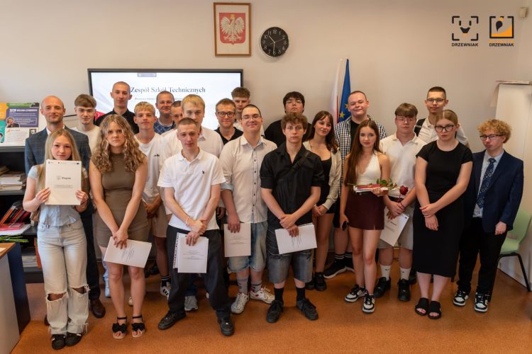 Pamiątkowe zdjęcie grupowe absolwentów szkoły. Uczniowie w galowych strojach pozują razem po odebraniu świadectw, które trzyma kilkoro z nich. W tle widać godło Polski, zegar, flagi oraz ekran z logiem ZST w Słupsku